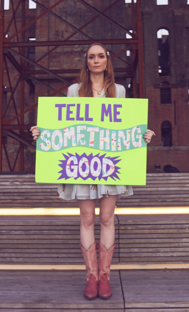 Singer Lauren Jaimes holding purple/green sign with the title of her new song, Tell Me Something Good.